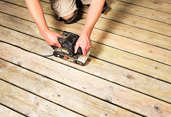 A person sanding a wooden deck with a belt sander, which is relevant as it visually represents the deck board sanding process involved in the described project.